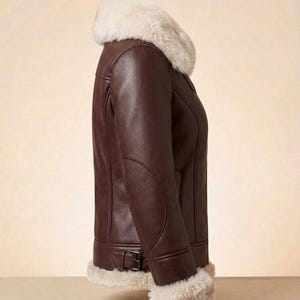 K&ouml;nnte beinhalten: Eine dunkelbraune Lederjacke mit einem dicken, cremefarbenen Kunstfellkragen und passenden Kunstfellb&uuml;ndchen an den &Auml;rmeln. Die Jacke verf&uuml;gt &uuml;ber einen seitlich verstellbaren Schnallenriemen am Saum und sichtbare Nahtdetails entlang des Rumpfes.