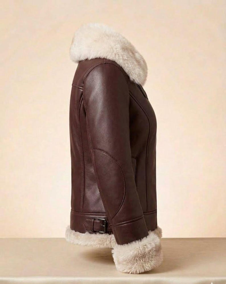 K&ouml;nnte beinhalten: Eine dunkelbraune Lederjacke mit einem dicken, cremefarbenen Kunstfellkragen und passenden Kunstfellb&uuml;ndchen an den &Auml;rmeln. Die Jacke verf&uuml;gt &uuml;ber einen seitlich verstellbaren Schnallenriemen am Saum und sichtbare Nahtdetails entlang des Rumpfes.