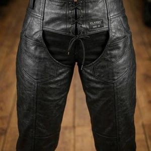 May include: Black leather pants with a lace-up front and a high waist. The pants have a classic design with pockets and a size 26 label. The leather appears textured and worn, giving them a vintage look.