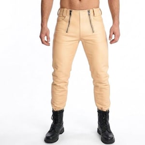 Può includere: Pantaloni in pelle color cuoio con due cerniere frontali e un design aderente. I pantaloni sono abbinati a stivali da combattimento neri. L'immagine è ripresa da una prospettiva frontale.