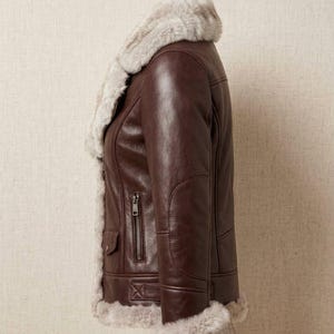 K&ouml;nnte beinhalten: Eine dunkelbraune Lederjacke mit einem dicken, hellbeigen Kunstfellfutter entlang des Kragens, der Vorder&ouml;ffnung und der &Auml;rmelb&uuml;ndchen. Die Jacke verf&uuml;gt &uuml;ber eine vertikale Rei&szlig;verschlusstasche an der Seite und eine kleine Pattentasche mit Knopf.
