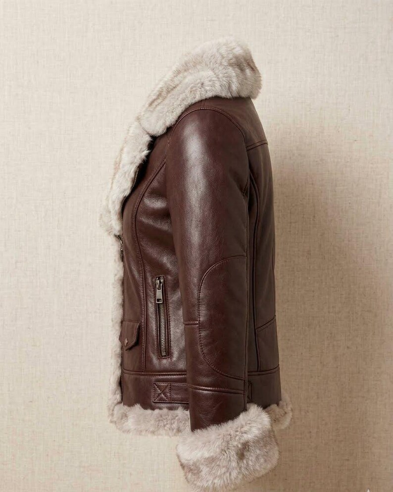 K&ouml;nnte beinhalten: Eine dunkelbraune Lederjacke mit einem dicken, hellbeigen Kunstfellfutter entlang des Kragens, der Vorder&ouml;ffnung und der &Auml;rmelb&uuml;ndchen. Die Jacke verf&uuml;gt &uuml;ber eine vertikale Rei&szlig;verschlusstasche an der Seite und eine kleine Pattentasche mit Knopf.