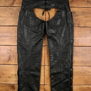 May include: Black leather chaps with a lace-up back and snap closures on the sides. The chaps have two back pockets and are designed with a classic fit. The leather appears worn, giving them a vintage look.