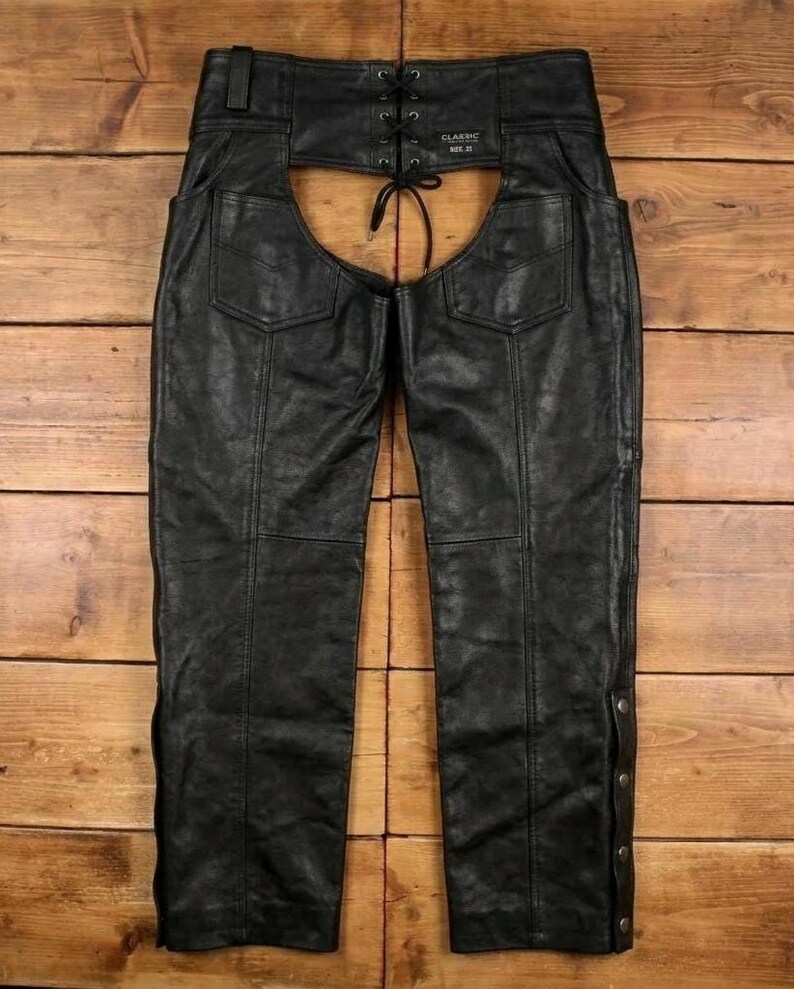 May include: Black leather chaps with a lace-up back and snap closures on the sides. The chaps have two back pockets and are designed with a classic fit. The leather appears worn, giving them a vintage look.