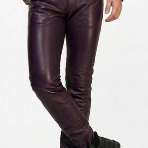 Puede incluir: Pantalones de cuero color ciruela con cinturón negro con tachuelas y botas negras. Los pantalones tienen un corte ajustado y un ligero brillo. Las botas son con cordones y suela robusta. El conjunto se completa con una chaqueta de cuero negra.