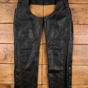 May include: Black leather chaps with a belt and buckle closure at the waist. The chaps have a classic design with a straight leg and are made of a durable material. The chaps have a row of silver-colored snaps along the side.
