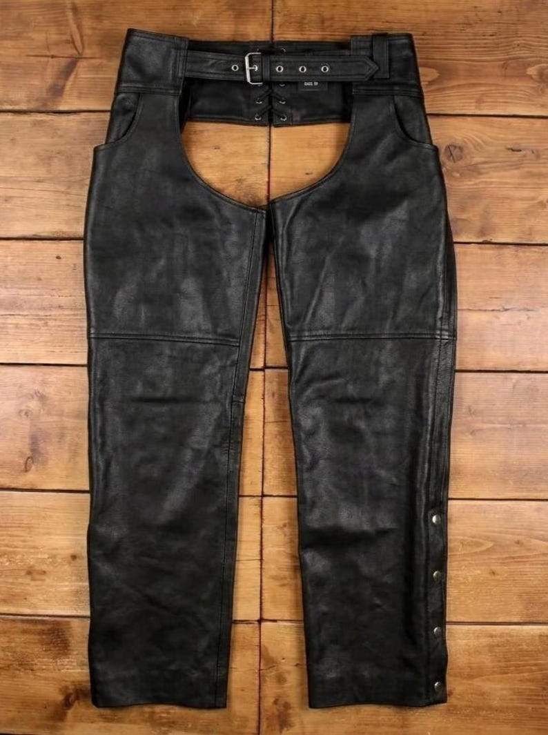May include: Black leather chaps with a belt and buckle closure at the waist. The chaps have a classic design with a straight leg and are made of a durable material. The chaps have a row of silver-colored snaps along the side.