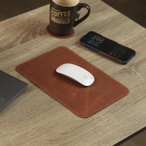 May include: A rectangular brown leather mouse pad with rounded corners, featuring a white wireless computer mouse centred on its surface. The mouse pad is positioned next to a dark grey laptop, a brown coffee mug with the text COFFEE MAKES EVERYTHING POSSIBLE, and a black smartphone.
