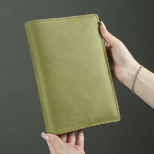 Könnte beinhalten: Ein rechteckiger Notizbuchumschlag aus olivgrünem Leder mit einer glatten, matten Oberfläche und sichtbaren Nähten entlang der Kanten. Der Umschlag hat ein schlichtes, minimalistisches Design mit einer einheitlichen Farbe und Struktur.