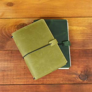 May include: Two leather journals stacked together. The top journal is light olive green, and the bottom journal is dark forest green. Both feature a smooth matte finish, visible stitching along the edges, and a black elastic band closure.