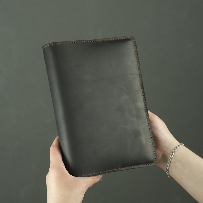 May include: A rectangular dark brown leather notebook cover with a smooth, matte finish and visible stitching along the edges. The cover is solid in color with a clean, minimalist design, featuring a vertical orientation and rounded corners.