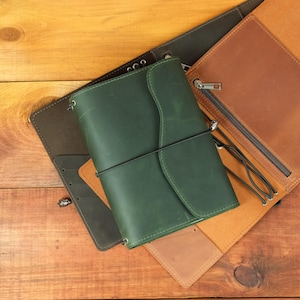 May include: A dark green leather journal cover with a flap closure and a black elastic band, layered over an open tan leather cover with a zippered pocket and a dark brown leather cover featuring multiple card slots.