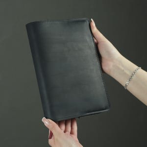 May include: A rectangular black notebook with a smooth, matte finish. The cover is solid black with visible stitching along the edges. A small, thin, dark red ribbon bookmark extends from the bottom edge of the notebook, providing a subtle contrast.