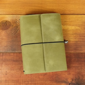 May include: A sage green leather journal cover with a smooth, matte finish. The rectangular cover features a vertical flap on the right side and is secured by a thin black elastic band that wraps horizontally around the center of the item.