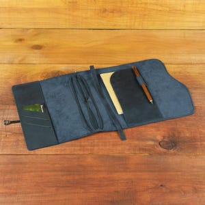 Op de afbeelding: Een marineblauwe lederen notitieboekhoes met meerdere binnenzakken, elastische banden voor het vastzetten van inserts en een houten pen in een speciale gleuf. Een kleine groene rechthoekige kaart zit in een zijvak.