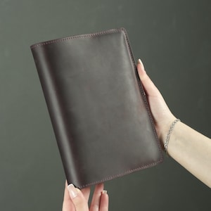 May include: A rectangular dark brown leather notebook cover with a smooth, matte finish and visible stitching along the edges. The cover is solid in color, featuring a clean, minimalist design with rounded corners and a simple, uniform construction.