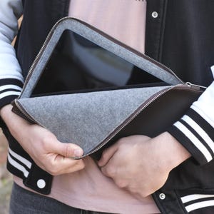 May include: A dark brown leather laptop sleeve featuring a silver metal zipper closure and a grey felt interior lining. The sleeve is partially open, revealing the soft grey felt material inside and the dark screen of a tablet or laptop device.