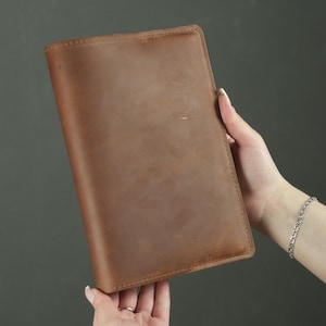 May include: A rectangular brown leather notebook cover with a smooth, matte finish and visible stitching along the top edge. The cover features a uniform tan-brown color with subtle natural variations in the texture of the leather surface.