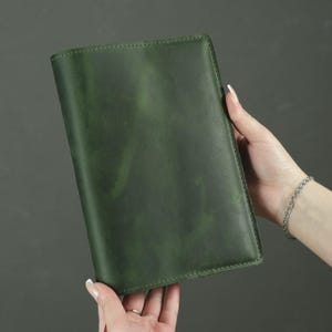May include: A rectangular notebook cover made of smooth, dark forest green leather. The cover features a matte finish with subtle tonal variations and visible stitching along the edges, providing a clean and structured appearance for a journal.