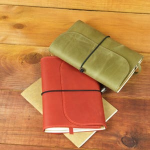 May include: Two leather journals, one in olive green and one in brick red, each featuring a smooth matte finish, a rounded front flap, and a thin black elastic band closure that wraps horizontally around the center of the covers.