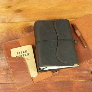 May include: A dark brown leather journal cover with a curved flap and black elastic closure. The interior features a tan leather pocket containing a Field Notes memo book and a matching leather pen loop holding a wooden pen.