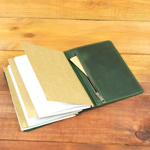 Puede incluir: Una funda de cuero verde oscuro para cuaderno con un bolsillo con cremallera en la solapa interior. El cuaderno contiene varias páginas de papel blanco y marrón claro, sujetas de forma segura dentro de la funda de cuero verde.