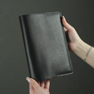May include: A rectangular black leather notebook cover with a smooth, matte finish and visible stitching along the edges. The cover is solid black, featuring a clean, minimalist design with rounded corners and a structured, durable appearance.
