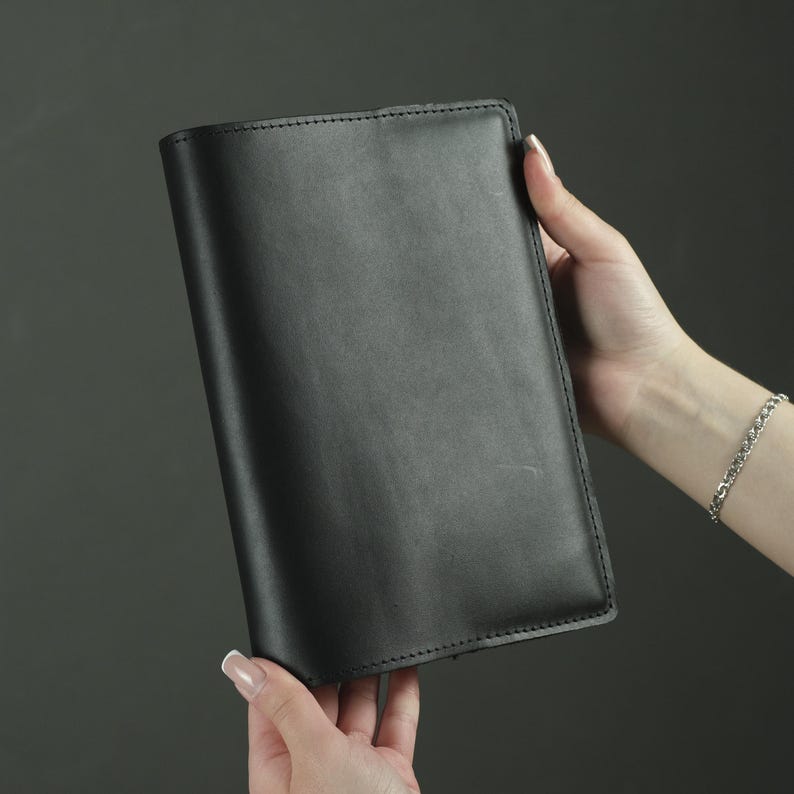 May include: A rectangular black leather notebook cover with a smooth, matte finish and visible stitching along the edges. The cover is solid black, featuring a clean, minimalist design with rounded corners and a structured, durable appearance.