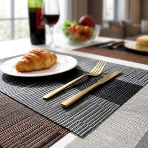 Puede incluir: Un mantel individual tejido rectangular con un patrón geométrico de rayas texturizadas en negro, gris y marrón. Un tenedor y un cuchillo de metal dorado están colocados lado a lado, junto a un plato blanco con un croissant dorado.