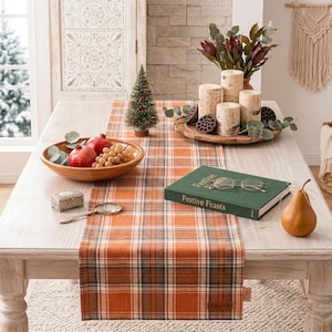 Puede incluir: Un camino de mesa a cuadros naranja, blanco y gris con una pequeña etiqueta de cuero. El camino se muestra sobre una mesa de madera con un cuenco de fruta, un pino en miniatura, un libro titulado Festive Feasts, velas y una lupa.