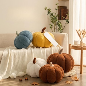 May include: A collection of five plush pumpkin-shaped pillows in blue, yellow, white, and two shades of brown. The pillows feature a textured, sherpa-like fabric with distinct vertical segments and a small brown stem on top of each pumpkin.