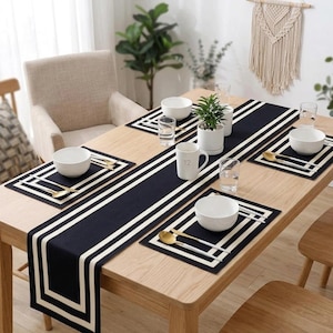May include: A black table runner and four matching rectangular placemats, each featuring a white border stripe design. The set is arranged on a light wood dining table with white ceramic bowls, gold-toned spoons, and clear glass tumblers.