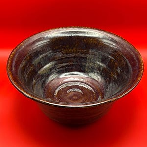 Può includere: Una ciotola in ceramica fatta a mano con una glassa marrone scuro e una finitura lucida. La ciotola ha un design ampio e aperto e una superficie strutturata. L'interno presenta sottili variazioni di colore. Questa ciotola decorativa è adatta per servire o esporre.