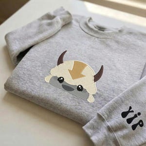 May include: A light gray crewneck sweatshirt featuring an embroidered graphic of a stylized creature with a tan arrow on its head, dark brown horns, and black eyes. The sleeve displays a black embroidered design of three vertical abstract shapes.
