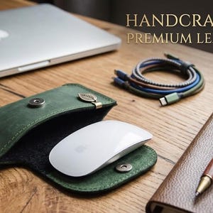 May include: A dark green leather sleeve with a snap button closure and a small metal leaf charm, containing a white computer mouse. The text HANDCRAFTED PREMIUM LEATHER is visible in the upper right corner of the image.