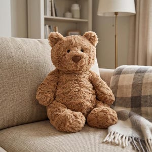 May include: A soft, brown teddy bear with textured fur sits on a beige sofa. The bear has a heart-shaped nose and small, rounded ears. A plaid blanket rests on the sofa next to the bear, creating a cosy, inviting scene.