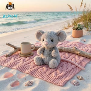 May include: A plush gray mouse toy with large ears and a pink nose sits on a pink cable-knit blanket on a sandy beach. A beige mug and a piece of driftwood are nearby. Seashells and a succulent in a terracotta pot complete the scene.