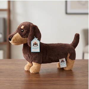 May include: A plush dachshund dog toy in brown and tan. The stuffed animal has a long body, floppy ears, and a small tail. It has a tag that says "Jellycat". The toy is on a wooden surface.