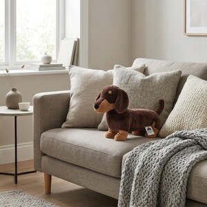 May include: A plush dachshund dog toy in brown and tan, positioned on a light gray sofa with matching pillows and a gray knitted throw. The dog toy has a tag attached. The room has a neutral color palette with a window in the background.