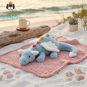 Op de afbeelding: Een lichtblauwe pluche draak met witte vleugels en kleurrijke accenten, liggend op een roze gebreide deken. De draak ligt op een zandstrand met een kopje en een vetplant op de achtergrond. Het Jellycat-logo staat linksboven.
