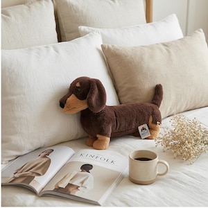 May include: A plush dachshund dog toy in shades of brown and tan rests on a bed with white and beige pillows. The dog has floppy ears and a long body. A magazine with the word "KINfolk" is open nearby, along with a mug of coffee.