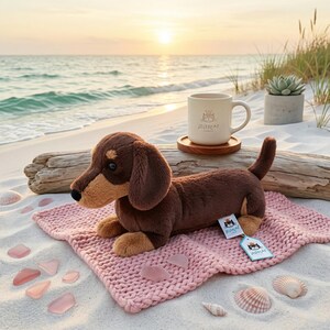 May include: A plush brown dachshund dog toy with tan accents, lying on a pink knitted blanket on a sandy beach. A white mug with a cat logo and a wooden coaster are nearby. The scene is set at sunset.