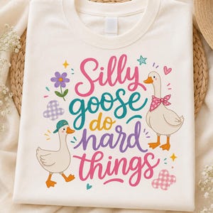 May include: A cream-colored t-shirt featuring a graphic print with the text "Silly goose do hard things" in pink, teal, yellow, and purple. The design includes two white geese, one wearing a teal baseball cap and one with a pink bandana.