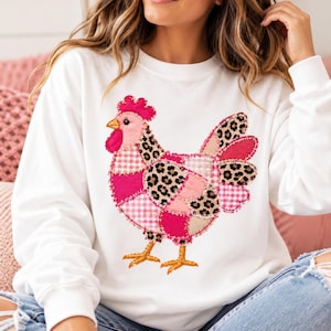 May include: A white long-sleeve sweatshirt featuring a large, central appliqué of a chicken. The chicken is constructed from various fabric patches in shades of pink, white gingham, and leopard print, with a bright pink comb and yellow feet.