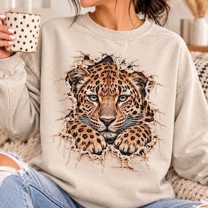 May include: A beige crewneck sweatshirt featuring a graphic print of a leopard face appearing to emerge through a torn fabric effect. The leopard has detailed black spots and golden fur. The sweatshirt is worn with blue denim jeans.