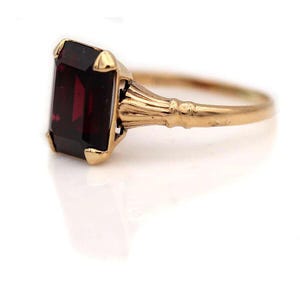 Könnte beinhalten: Ein goldener Ring mit einem rechteckigen, tiefroten Edelstein in einer vierzackigen Fassung. Die Ringschiene weist ein geripptes, kanneliertes Design auf, das von der Fassung ausgeht und eine polierte metallische Oberfläche sowie eine klassische Form zeigt.