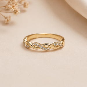 Op de afbeelding: Een goudkleurige ring met een gevlochten ontwerp. De ring is versierd met kleine, heldere edelstenen die langs het gevlochten patroon zijn geplaatst. De ring wordt gepresenteerd op een neutrale achtergrond, met zachte verlichting.