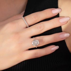 May include: A hand displaying two diamond rings. The main ring has a rectangular-cut diamond, complemented by smaller stones. The second ring is a thin band set with small diamonds. The hand has a light skin tone, set against a black backdrop.