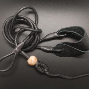 May include: A black leather cord lanyard featuring a textured leather wrist loop, a smooth round beige bead, and a black braided knot detail. The cord is coiled and secured with dark grey heat shrink tubing at the connection points.