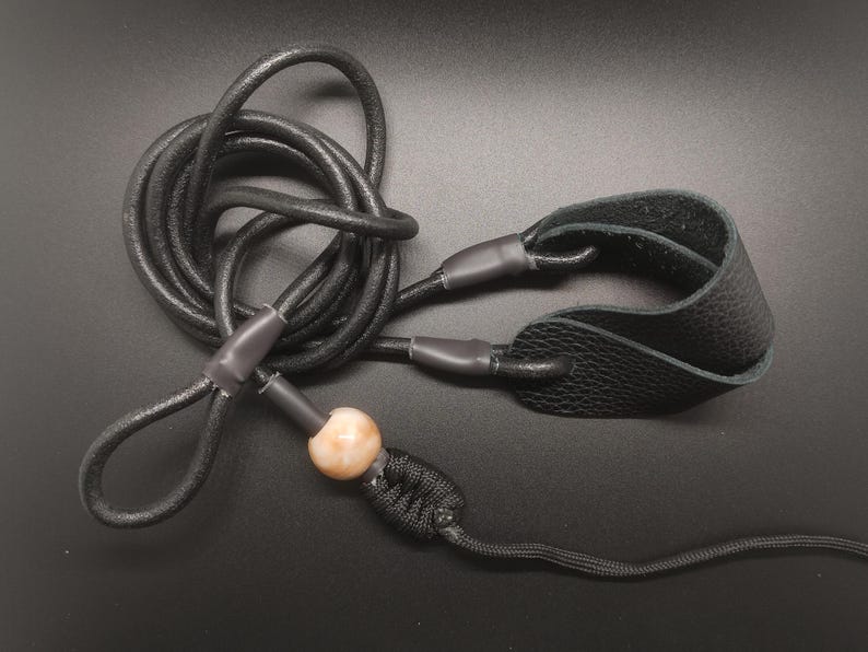 May include: A black leather cord lanyard featuring a textured leather wrist loop, a smooth round beige bead, and a black braided knot detail. The cord is coiled and secured with dark grey heat shrink tubing at the connection points.